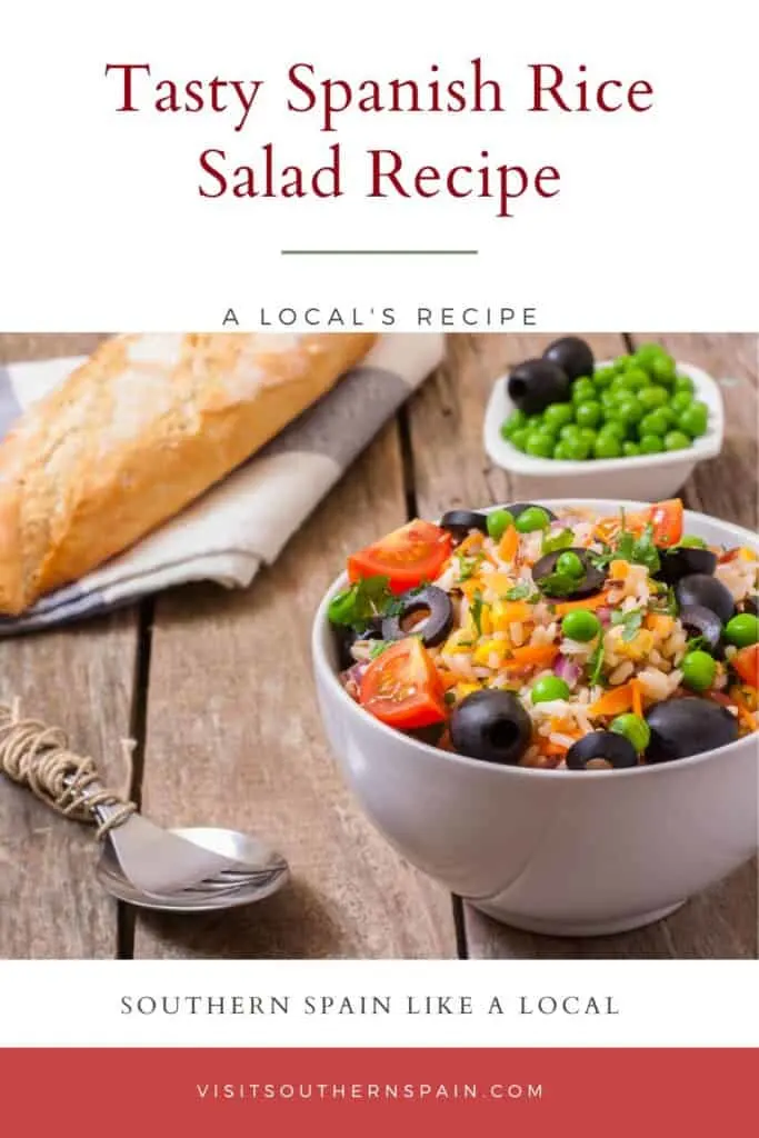 a pin with a Spanish rice salad with olives, tomatoes and vegetables in a bowl with bread in the background.