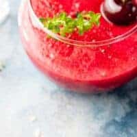 cherry gazpacho in a glass bowl decorated with a whole cherry and fresh herbs