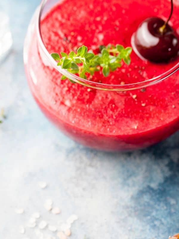 cherry gazpacho in a glass bowl decorated with a whole cherry and fresh herbs - Easy Spanish Cherry Gazpacho Recipe
