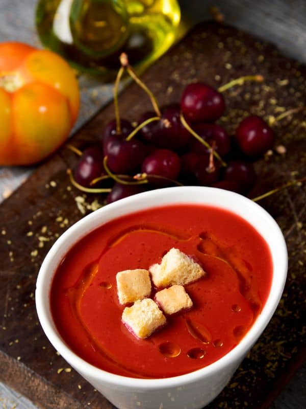 cherry gazpacho served in a white bowl with croutons and olive oil next to fresh cherries and tomato