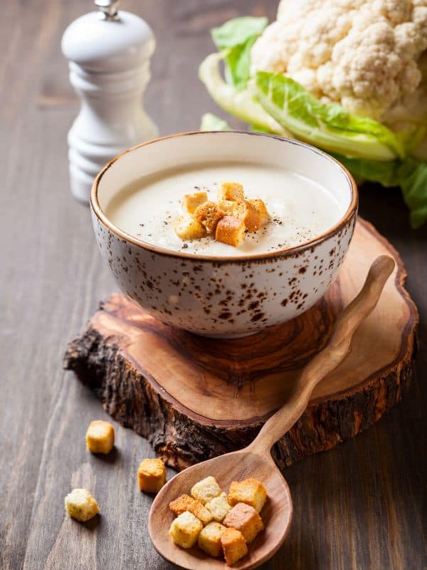 cold cauliflower soup served in a bowl with croutons, on a wooden board with cauliflower next to it