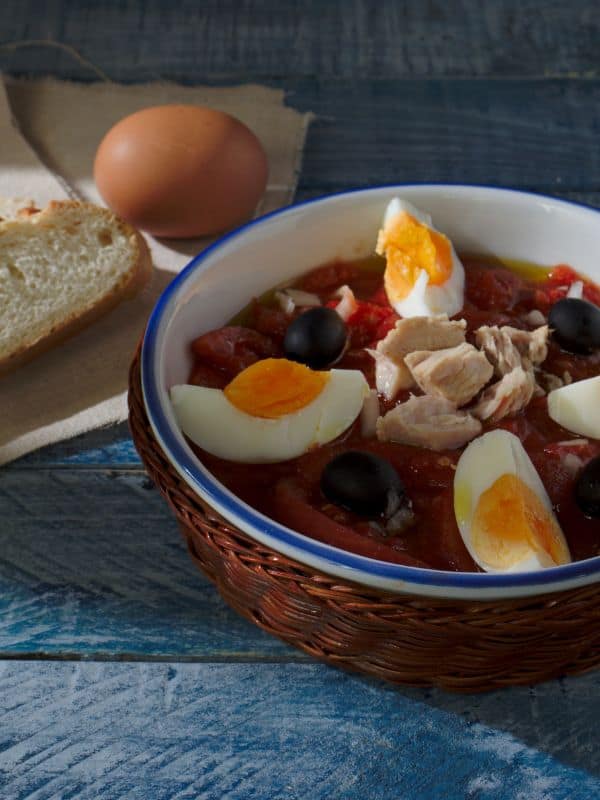 murcia salad with boiled eggs, tomatoes, olives and tuna with a slice of bread and an egg next to the bowl