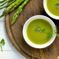 2 bowls of cold asparagus soup on a wooden board next to fresh asparagus.