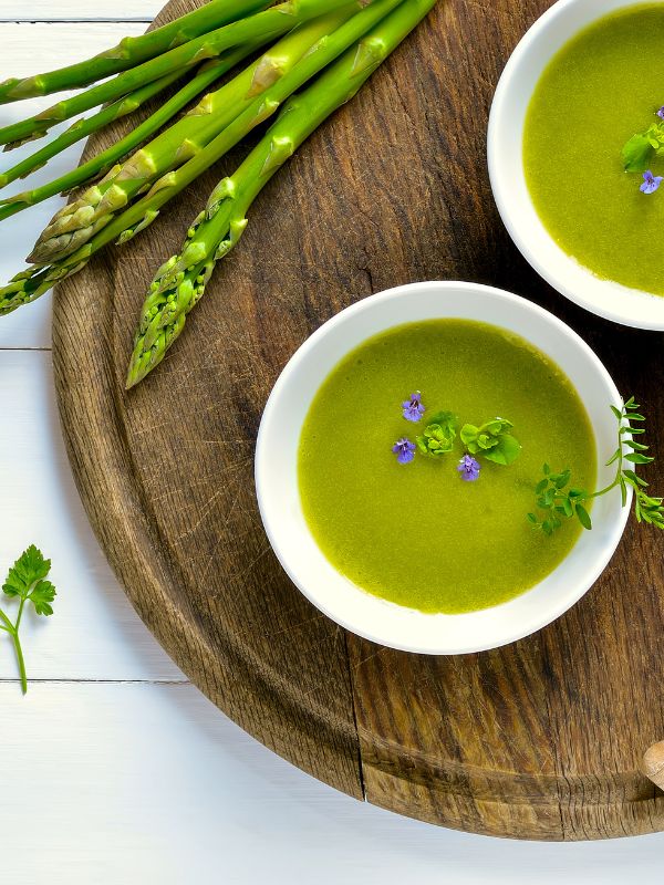 2 bowls of cold asparagus soup on a wooden board next to fresh asparagus. - Spanish Cold Asparagus Soup Recipe