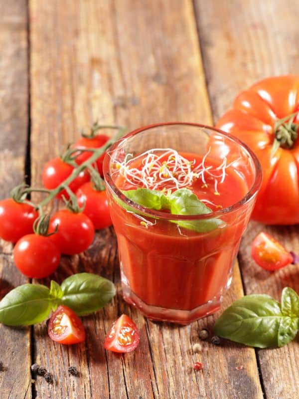 a glass of gazpacho shot with fresh tomatoes next to it on a wooden table.