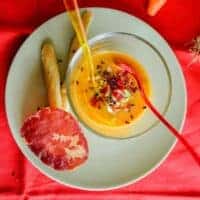 yellow tomato gazpacho in a glass bowl with carrot and bread sticks next to it.