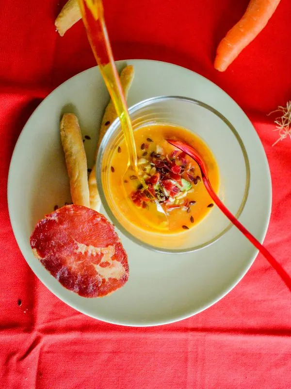 yellow tomato gazpacho in a glass bowl with carrot and bread sticks next to it.