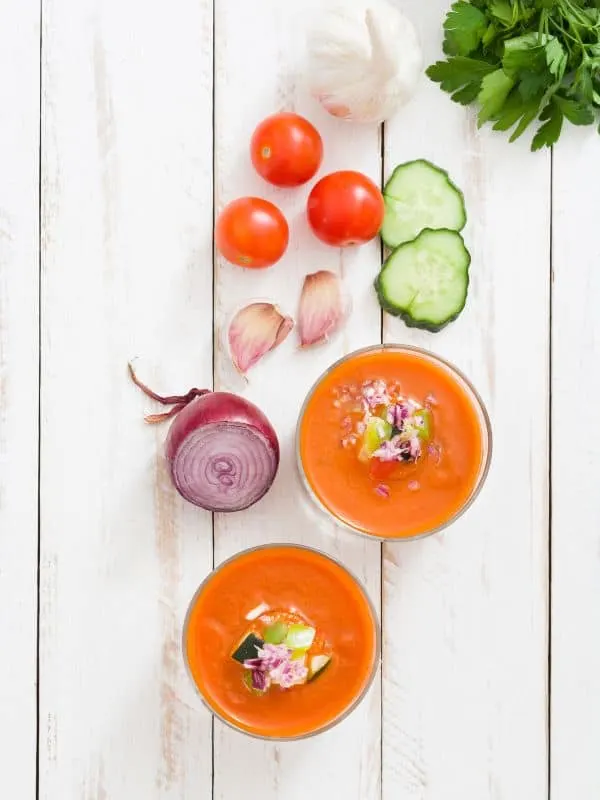 tomato basil gazpacho in 2 bowls with fresh vegetables next to them