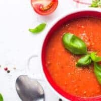 avocado tomato soup in a bowl with half of tomato next to it and decorated with basil