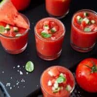 Gazpacho shot in small glasses with watermelon and tomatoes next to them