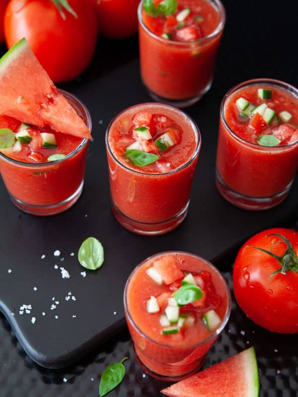 Watermelon Tomato Gazpacho in small glasses with watermelon and tomatoes next to them, a classic among spanish recipes