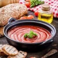 best tomato gazpacho in a black bowl with bread, tomatoes and olive oil next to it on a wooden surface.