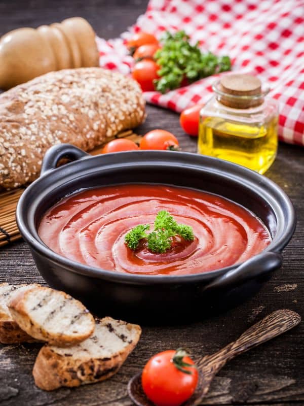 best tomato gazpacho in a black bowl with bread, tomatoes and olive oil next to it on a wooden surface.