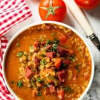 a big bowl of spanish lentil soup with chorizo next to 2 tomatoes.