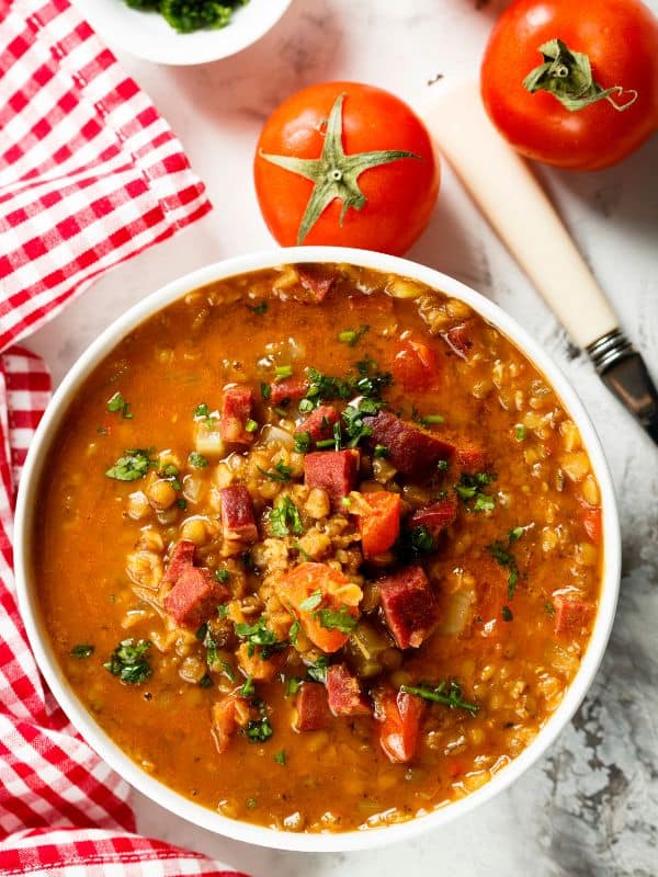 a big bowl of spanish lentil soup with chorizo next to 2 tomatoes.