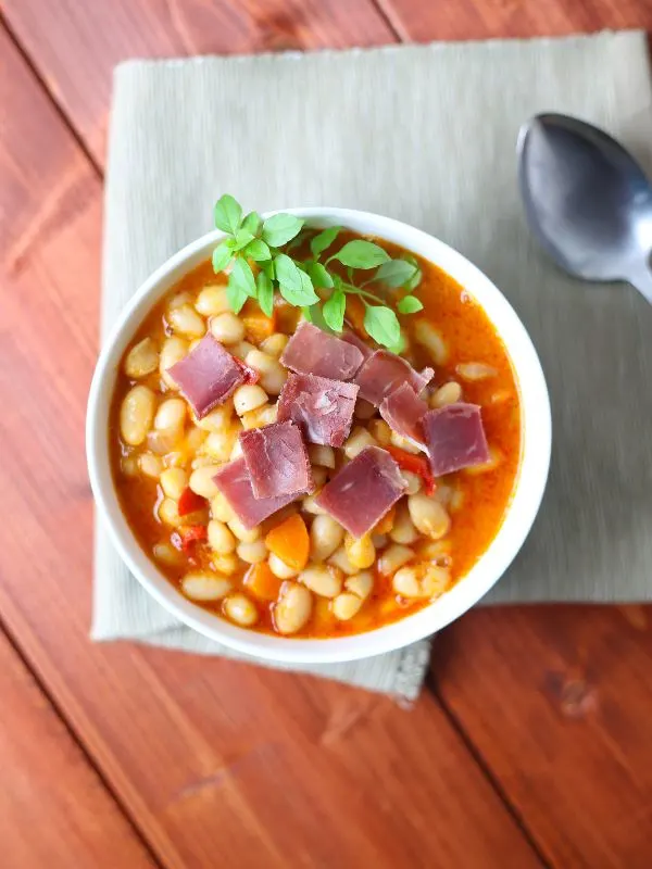 a pin with a bowl of Spanish bacon bean soup on a wooden table.