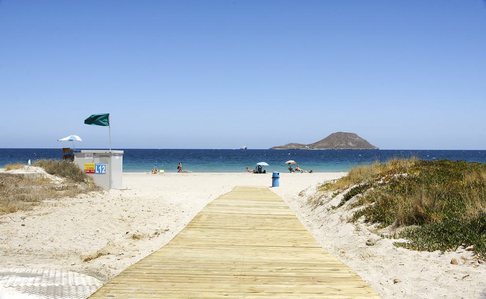 a wooden walkway on the beach with a sign, grass on the side, with people sun bathing on the beach, and an island with a mountain on the background