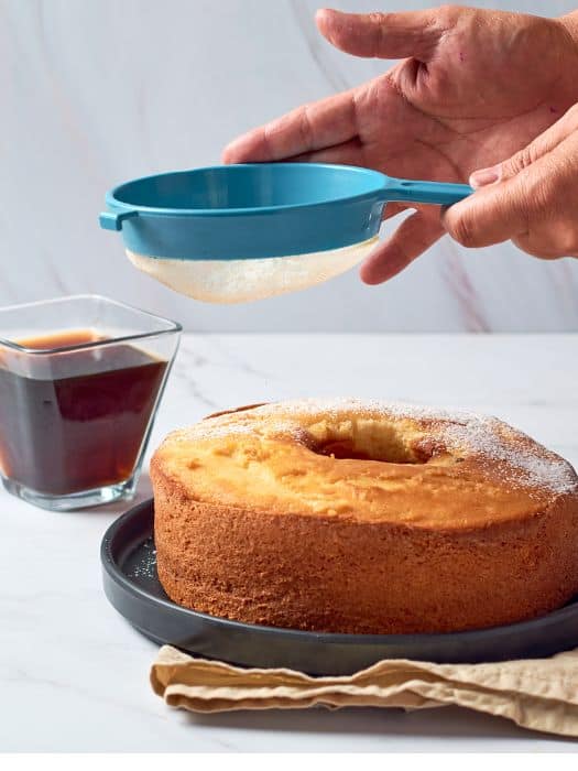 churros cake being dusted with sugar next to a cup of tea