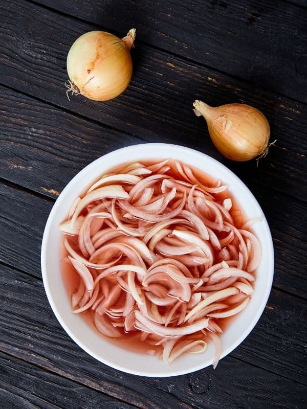 spanish pickled onions in a bowl on a dark wooden table next to 2 white onions